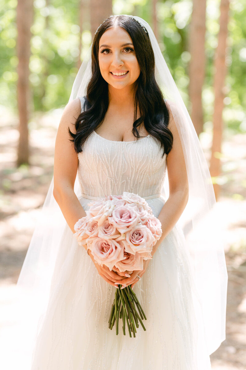 Bride holding a blush rose bouquet in a sunlit woodland setting