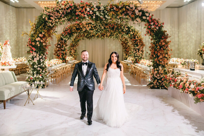 Bride and groom walking beneath a large floral ceremony arch