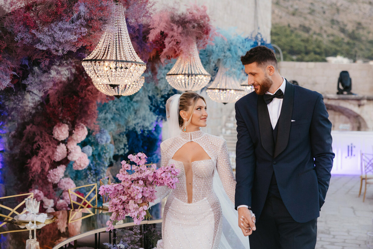 Bride and groom walking hand in hand beneath crystal chandeliers and dramatic pink and blue floral installations